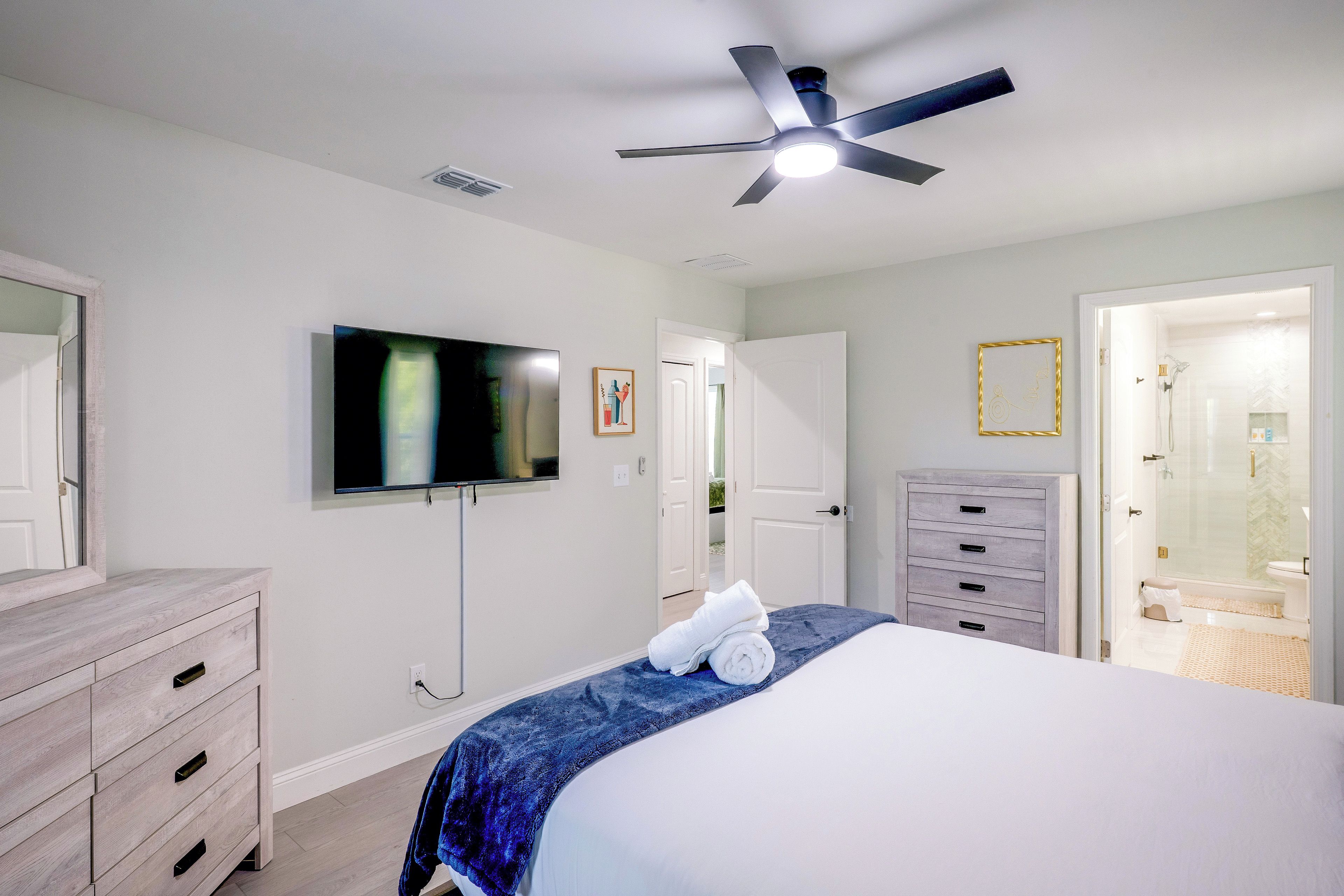 A bedroom with a bed, a television, a ceiling fan, a dresser, and a bathroom visible through an open door.