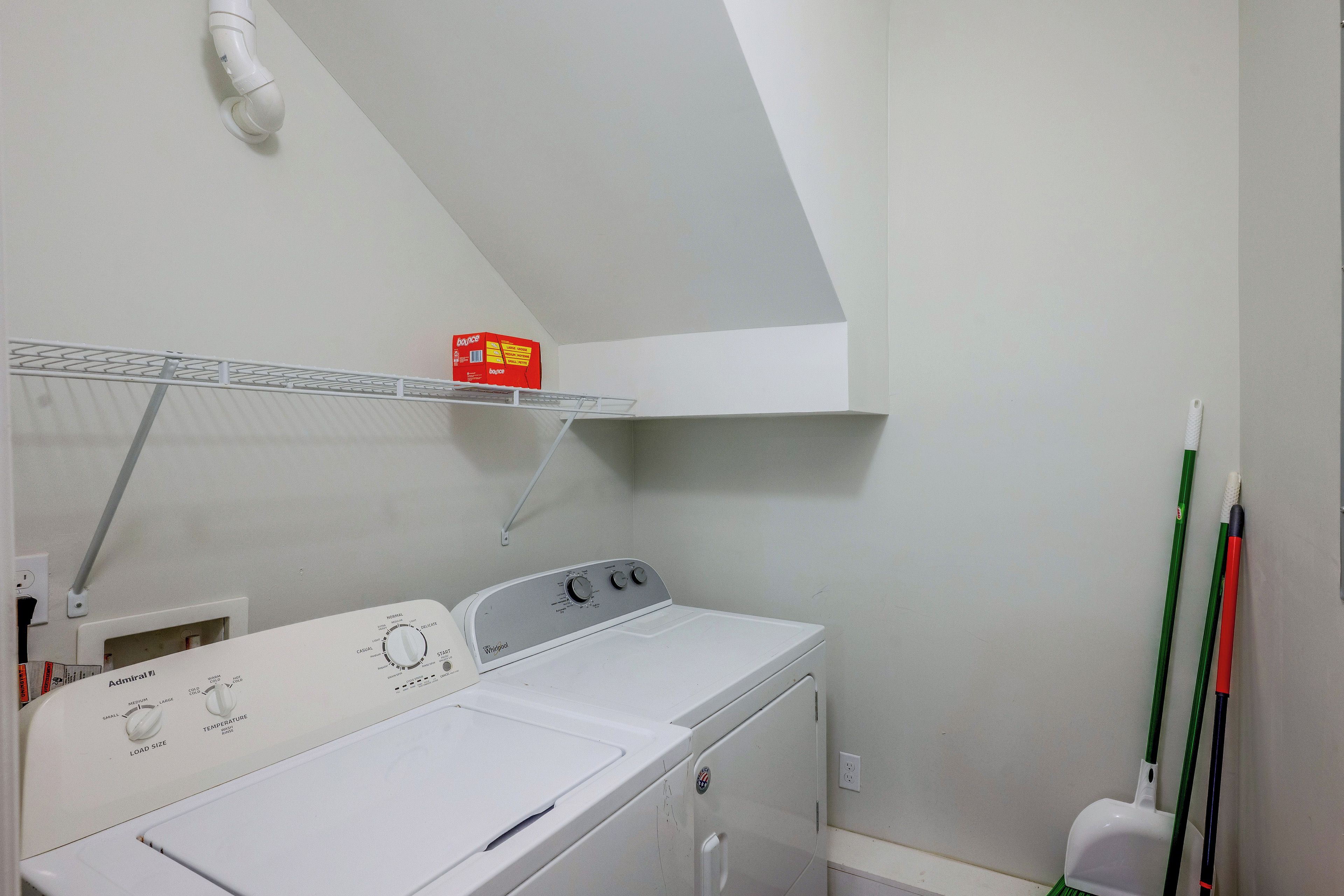 A laundry room with a front-loading washing machine, a shelf with a box, and a mop on the right.