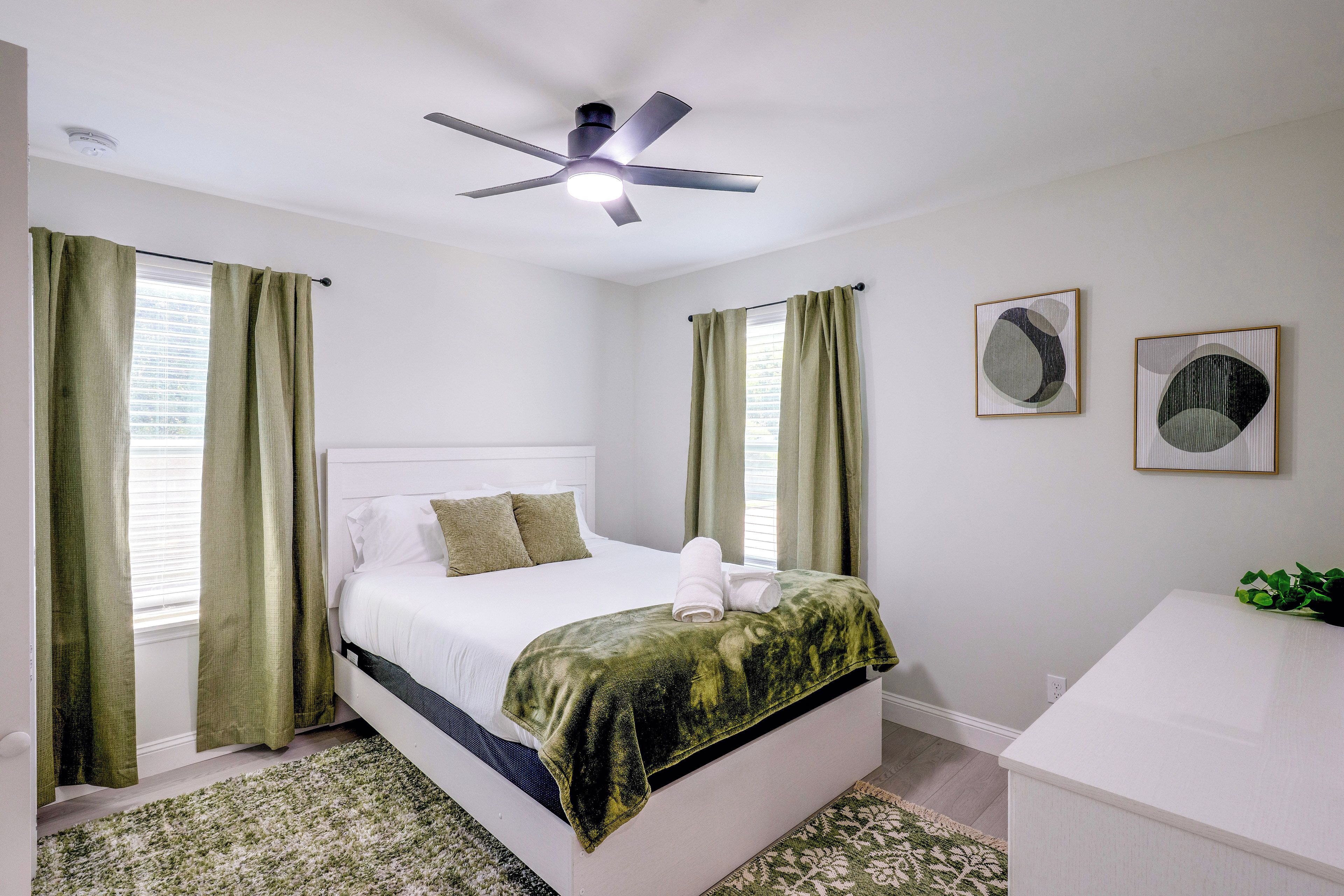 Ein Schlafzimmer mit einem Bett, einem Deckenventilator, einem Fenster mit Vorhängen, einem Nachttisch und einem an der Wand hängenden Kunstwerk.