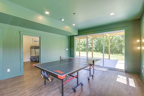 A ping pong table with wheels in a room with wooden flooring and a view of a garden through a glass door. - Brand New, Hot Tub & Porch, Near Nolin Lake! (Clarkson)