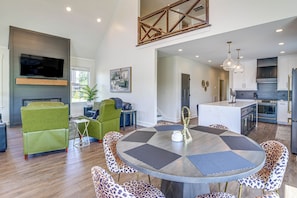 A modern kitchen with a central island, a dining area with a round table, and a living room with green sofas. - Brand New, Hot Tub & Porch, Near Nolin Lake! (Clarkson)