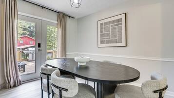 A dining area with a round table, four chairs, a light fixture, a door with curtains, and a framed abstract artwork on the wall.