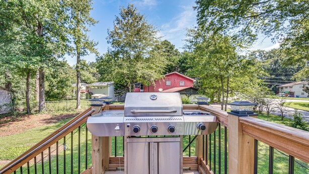 A barbecue grill on a wooden deck with a view of a red house and trees in the background.