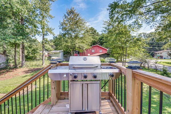A barbecue grill on a wooden deck with a view of a red house and trees in the background.