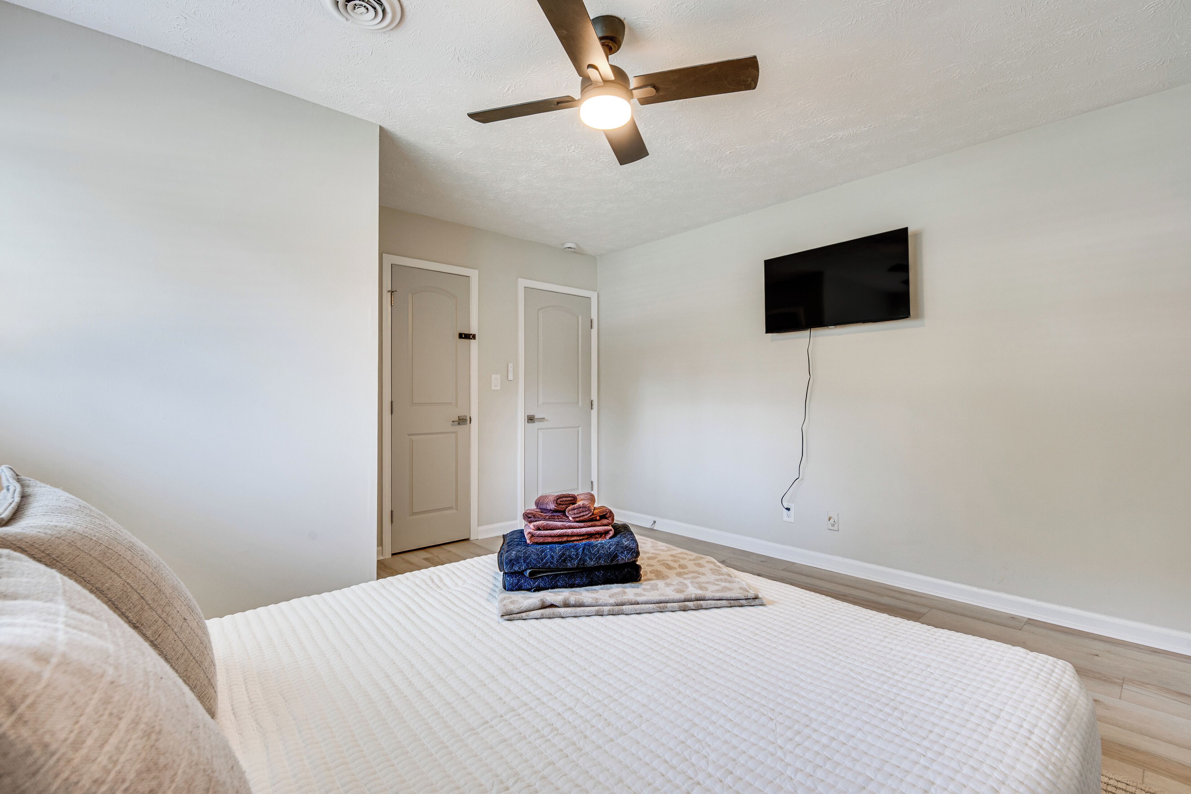 Ein Schlafzimmer mit einem Bett, einem Deckenventilator, einem an der Wand befestigten Fernseher und einer Tür zu einem anderen Raum.