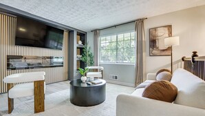 Una sala de estar moderna con televisor de pantalla plana, una mesa de centro redonda, un sofá y un amplio ventanal con cortinas.