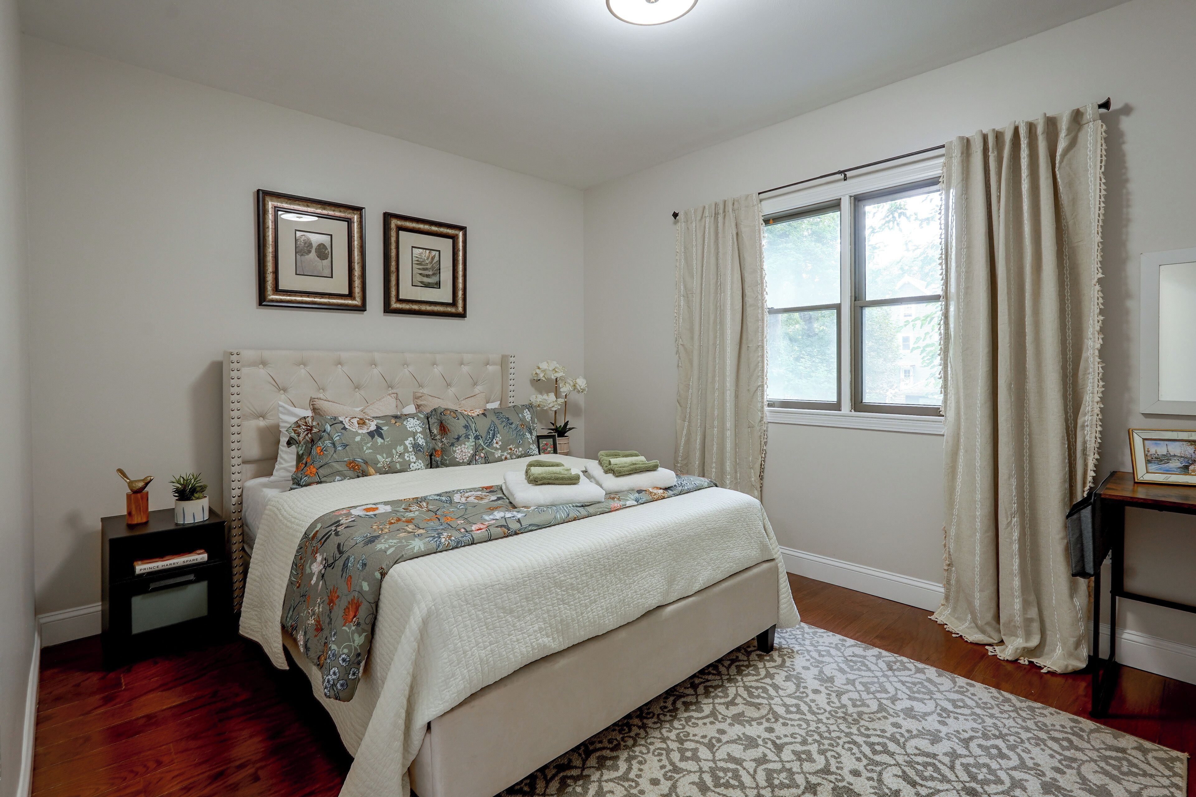 A bedroom with a large bed, a bedside table, a nightstand, a window with curtains, and two framed pictures on the wall.
