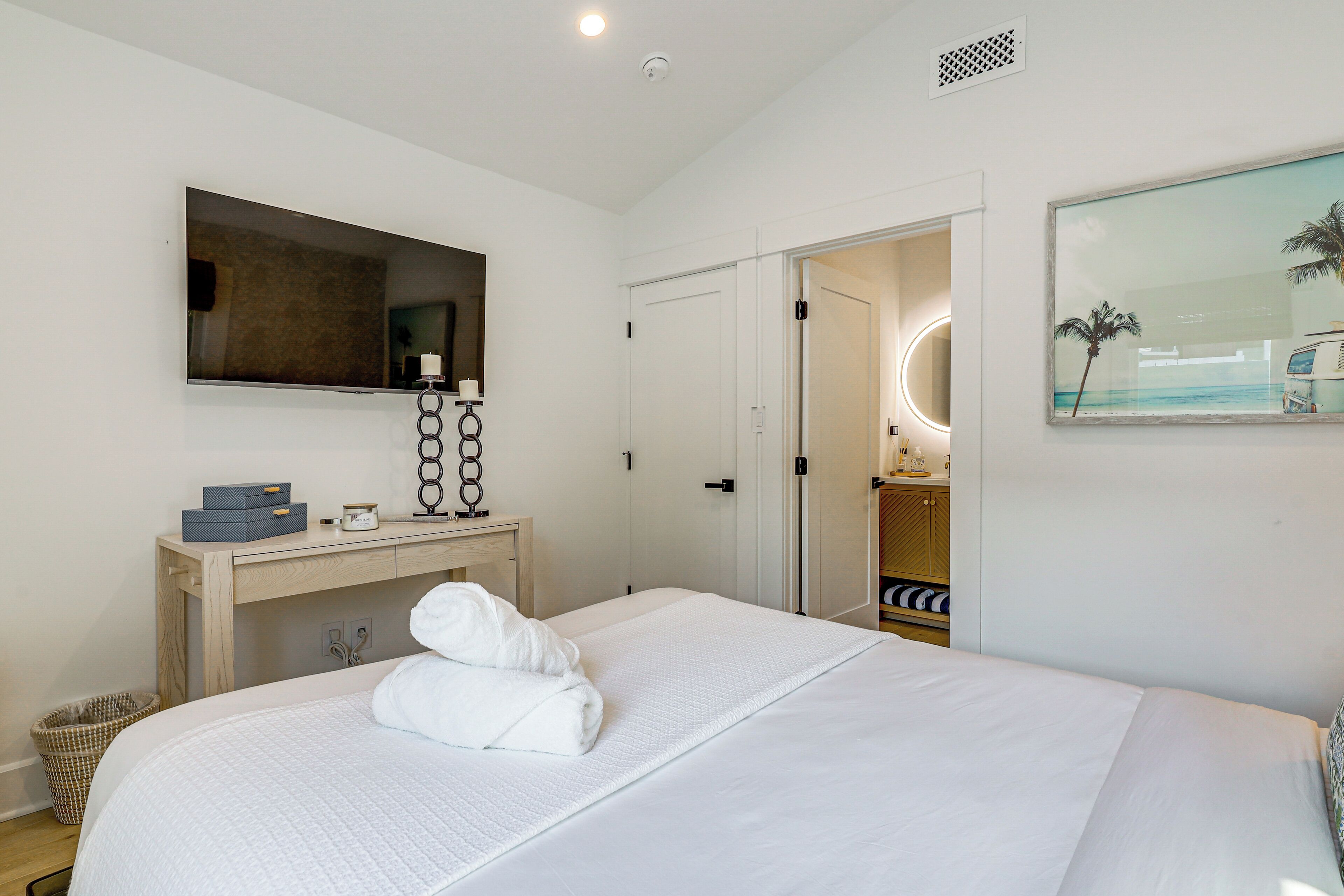 Habitación de hotel con cama, mesita de noche, espejo, cuadro y puerta que da a un baño.