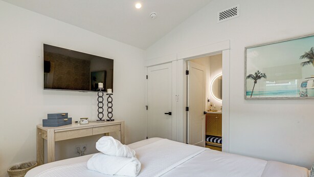 Habitación de hotel con cama, mesita de noche, espejo, cuadro y puerta que da a un baño.