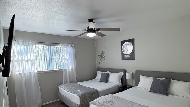 A hotel room with two beds, a ceiling fan, a window with curtains, a wall-mounted TV, and a framed picture. - Cozy 2-bedroom townhome by Galleria Houston with Pool & BBQ (Houston)