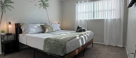 A bedroom with a bed, a ceiling fan, a nightstand with a lamp, a window with curtains, and a wall with a palm tree mural.