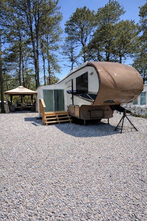 A recreational vehicle (RV) parked on a gravel surface with a covered outdoor seating area and a canopy structure nearby. - Trailer at the Peters Pond on beautiful Cape Cod! (Sandwich)