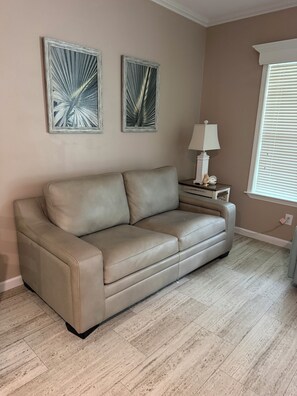 Un canapé en cuir beige est placé contre un mur, où sont accrochées deux peintures abstraites encadrées. On y trouve également une petite table de chevet avec une lampe, et une fenêtre avec des stores.