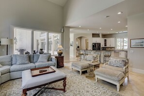 A spacious living room with a large window, a dining area with a kitchen in the background, and a cozy seating area with a patterned rug. - 9 Beach Lagoon | Lagoon Luxury with Infinity Pool | Walk to the Beach (Hilton Head Island)