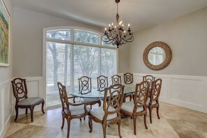 Ein Esszimmer mit einem runden Glastisch, der von verzierten Stühlen, einer Chandelierleuchtre und einem großen Fenster mit Jalousien umgeben ist.