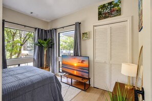 A hotel room with a bed, a television displaying a sunset, a window with curtains, and a door with louvered shutters. - 82 South Sea Pines: Artful Midcentry Modern Living in the Heart of Sea Pines (Hilton Head Island)