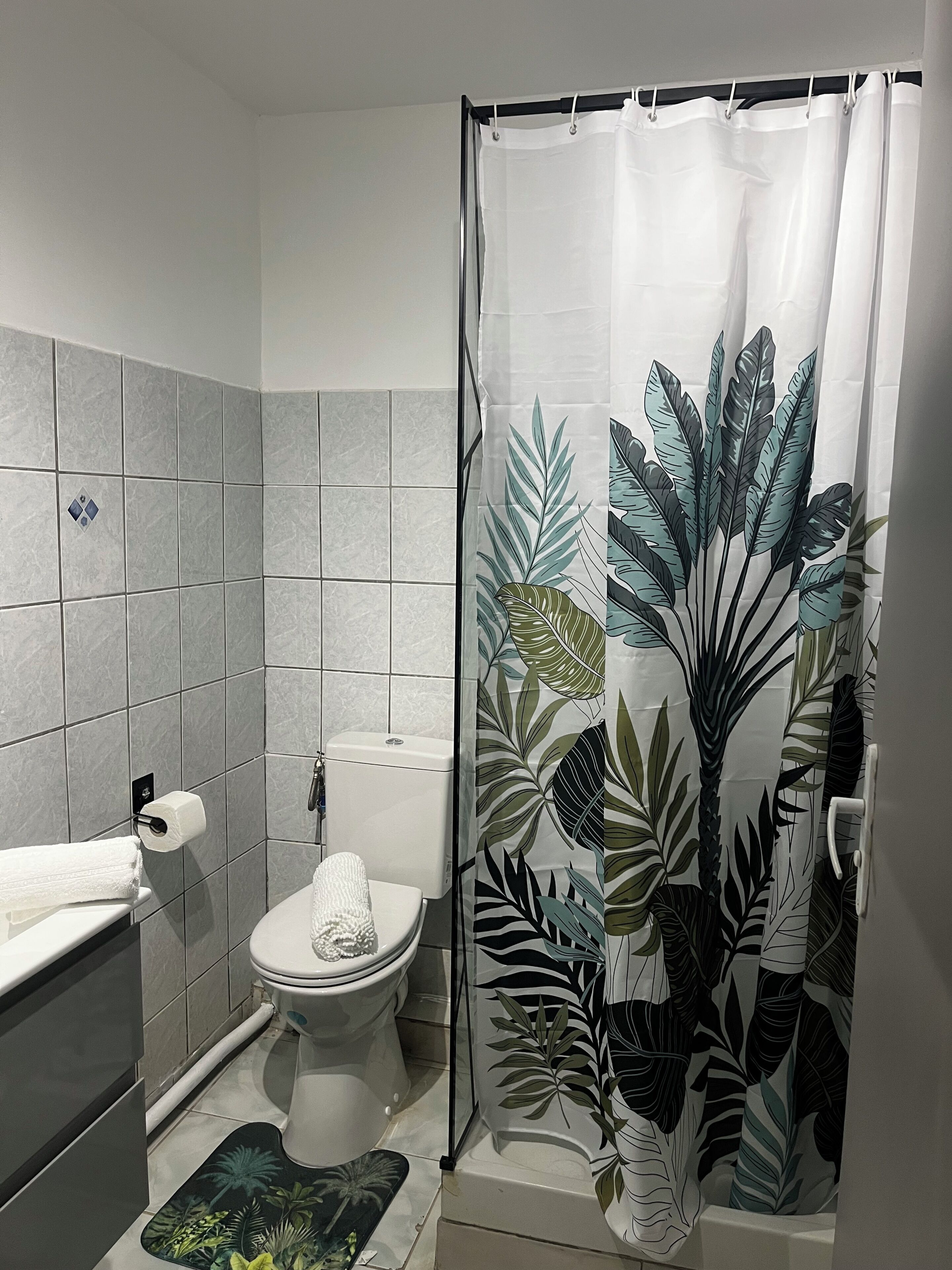 Ein Badezimmer mit Toilette, Handtuchhalter, einer Duschvorhang mit Blattmuster und einer Matte mit tropischem Design.