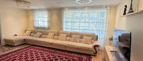 Un salon comprenant un canapé d’angle, un grand tapis rouge à motifs, un téléviseur à écran plat et des objets décoratifs accrochés au mur.