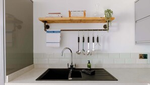 Eine Küche mit einem schwarzen Spülbecken, einem Holzregal über der Arbeitsplatte und verschiedenen Küchenutensilien, die an einem Gestell hängen.