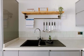 A kitchen with a black sink, a wooden shelf above the countertop, and various kitchen utensils hanging on a rack. - The Travelers Rest - Central Frome (Somerset)