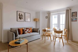 A modern living room with a grey sofa, a small round table, and a dining area with a white table and wooden chairs. - The Travelers Rest - Central Frome (Somerset)