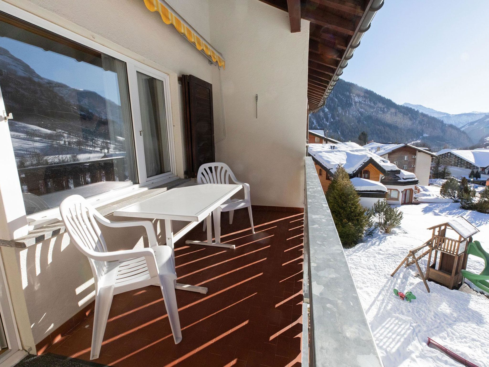 Un balcon avec une table et des chaises, donnant sur un village enneigé, avec des maisons et des montagnes en arrière-plan.