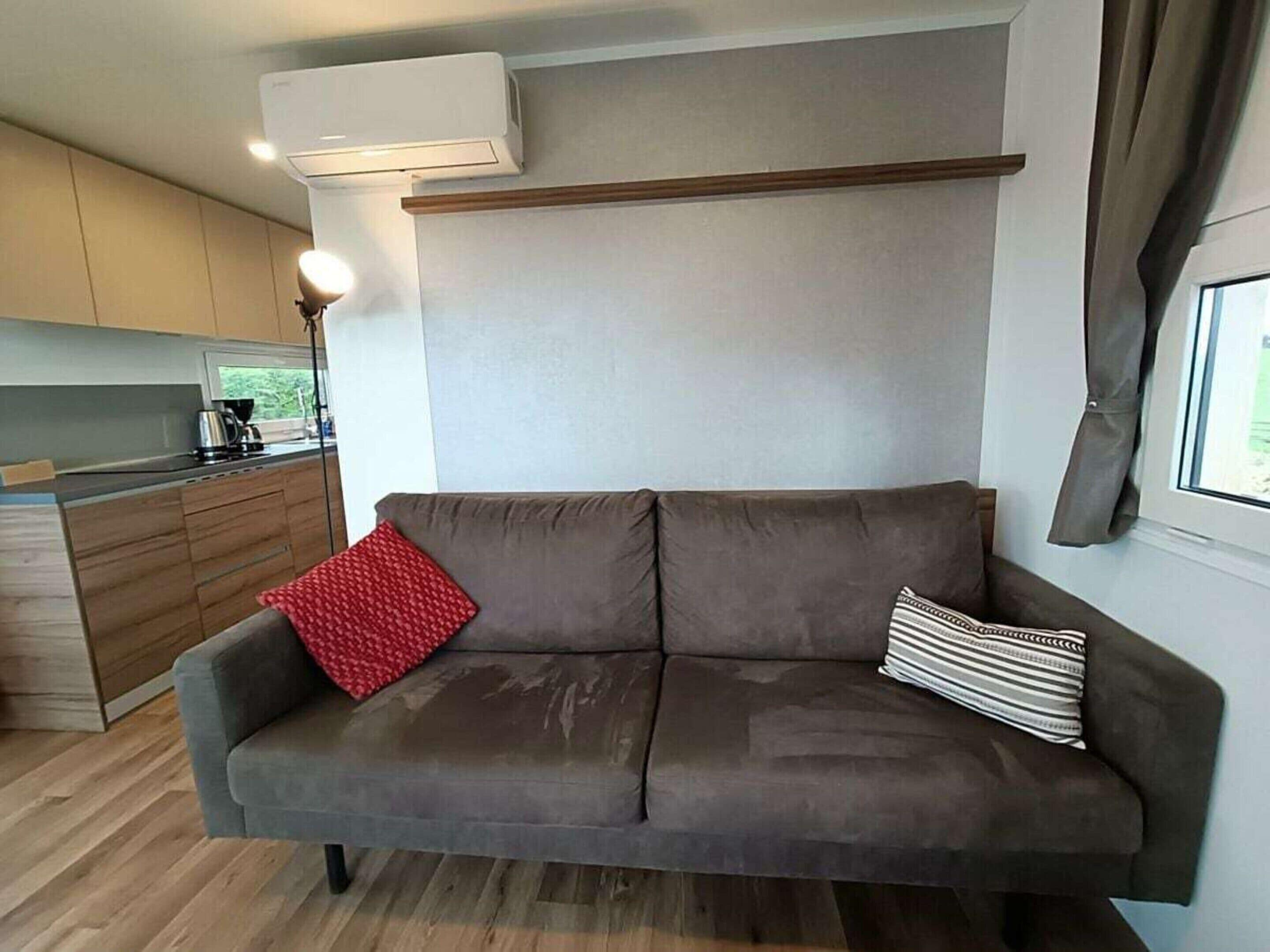 Een moderne woonkamer met een bank, een rood kussen en een gestreept kussen. Er is een keuken met houten kasten en een raam.
