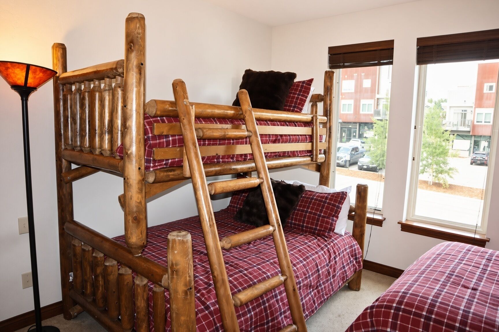 Een stapelbed met een ladder, een rode geruite sprei, en een raam met uitzicht op gebouwen en auto's.