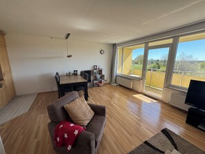 A living room with a sofa, a dining table, and a TV. There is a large window with a view of the outdoors. - Heimathafen Harz Vacation Rental – Hohegeiß, Pool & Sauna (Braunlage)