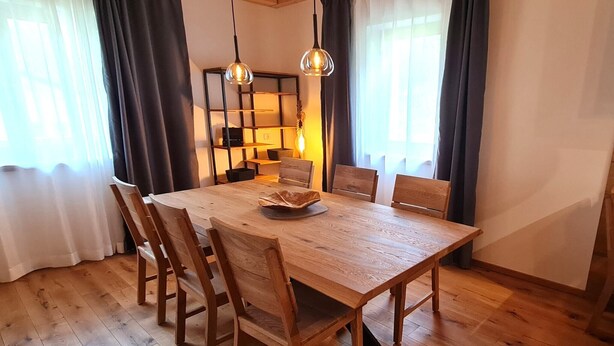 A dining area with a wooden table, six chairs, two pendant lights, and a bookshelf in the background.
