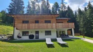 A two-story wooden chalet with a balcony, a covered patio area, and a lawn with two lounge chairs.