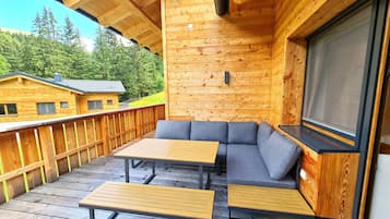A wooden deck with a sofa and two tables, a window, and a view of a forested area.