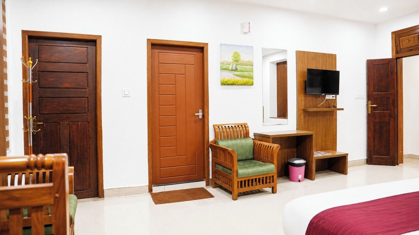 A hotel room with a wooden door, a green upholstered chair, a television on a wooden stand, and a bed with a red bedspread.