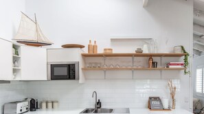 A modern kitchen with white cabinetry, a microwave, a toaster, and a sink. There are wooden shelves above the counter holding glassware and books. - The Lodge, Noosa - Stylish Retreat with Pool (Noosa Heads)