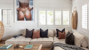 A living room with a beige sofa, multiple cushions, a woven basket, a framed picture, and a window with blinds. - The Lodge, Noosa - Stylish Retreat with Pool (Noosa Heads)