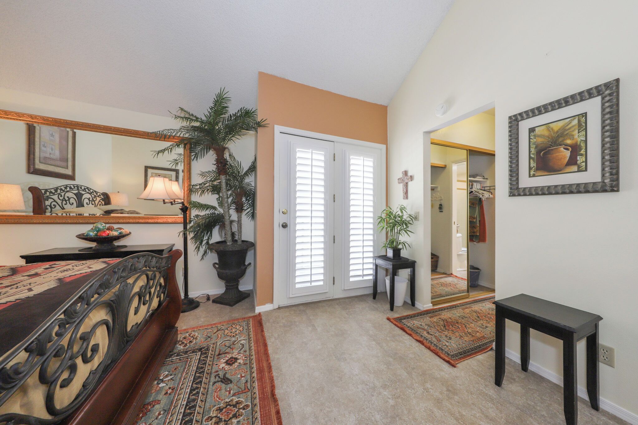 A hotel room with a bed, a desk, a lamp, a plant, a mirror, and a door leading to another room.
