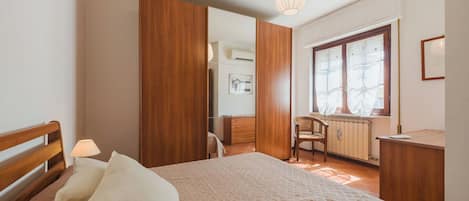 A bedroom with a bed, bedside table, lamp, wooden wardrobe, and a window with curtains.