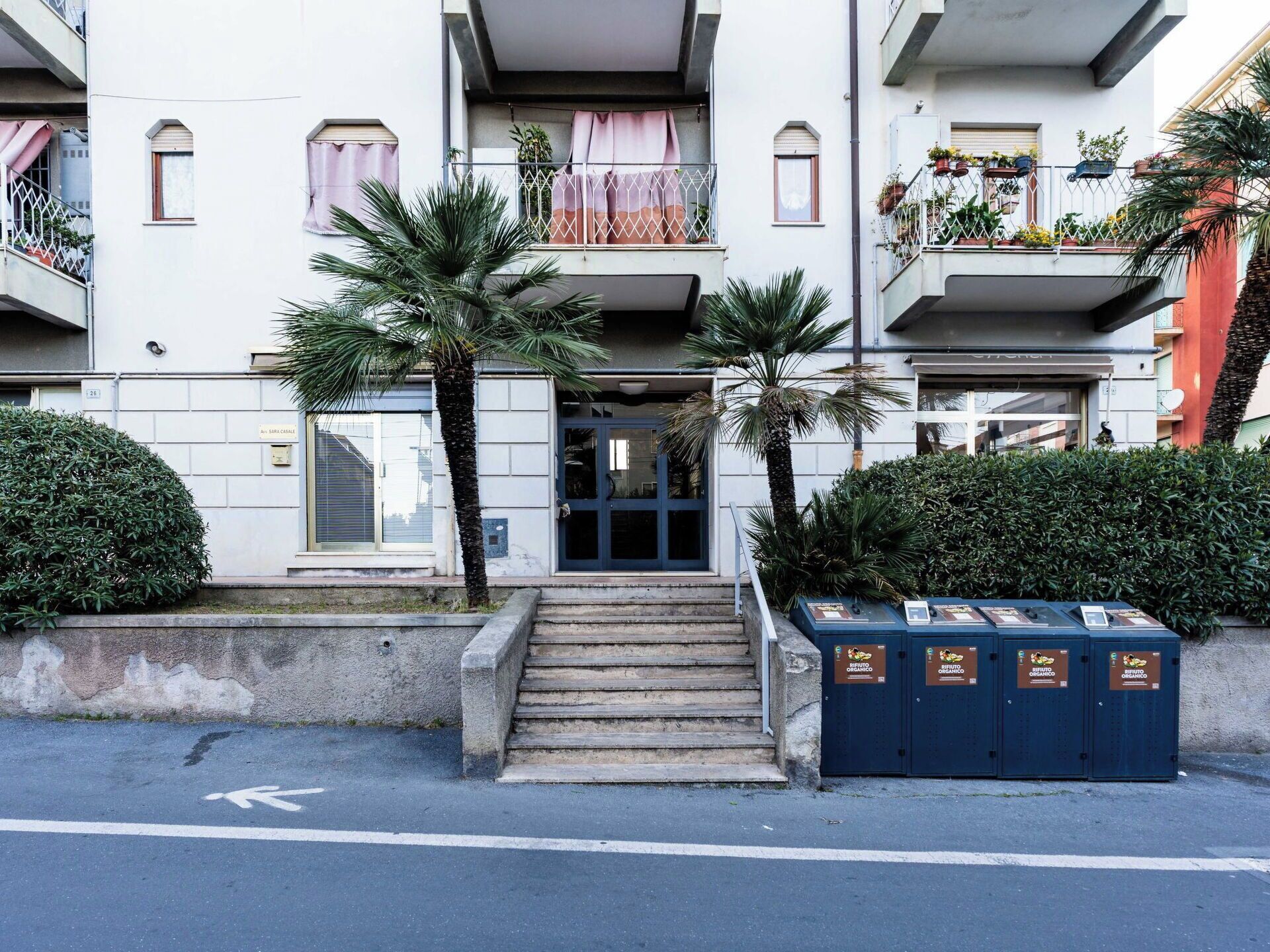 A multi-story residential building with a central entrance, a staircase leading to the entrance, and a row of balconies with potted plants.