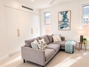 A modern living room with a grey sofa, a large abstract painting, a lamp, and a view of a brick building through the window. - Palm Springs Family Home – Gym, Garage, Pets, 10 minutes drive from Manly beach (North Balgowlah)