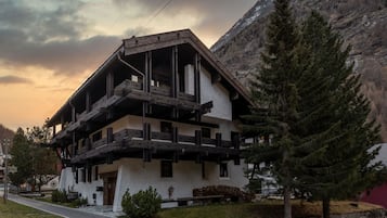 Ein mehrstöckiges Gebäude mit einer hölzernen Balkonkonstruktion, umgeben von Bäumen und einem Berg im Hintergrund.