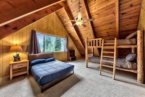 A cozy attic bedroom with a bunk bed, a single bed, a window with a view of trees, and a ceiling fan. - Tahoe River Chalet 2782b 4 Bedroom Home (South Lake Tahoe)