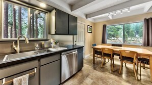 A modern kitchen with a built-in dishwasher, a dining area with a wooden table and chairs, and large windows offering a view of the forest. - Tahoe River Chalet 2782b 4 Bedroom Home (South Lake Tahoe)