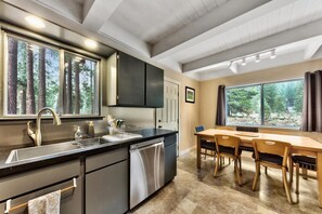 A modern kitchen with a built-in dishwasher, a dining area with a wooden table and chairs, and large windows offering a view of the forest. - Tahoe River Chalet 2782b 4 Bedroom Home (South Lake Tahoe)