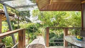 A covered outdoor area with a wooden deck, a table with a bowl of fruit, and a view of lush greenery.
