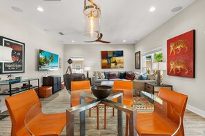A modern living room with a dining area, featuring a large flat-screen TV, a ceiling fan, and a vibrant abstract painting. - Arlington Park Oasis 4 Bedroom Home (Sarasota)