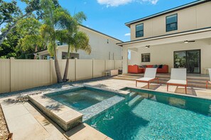 A swimming pool with a tiled deck, a covered seating area with a sofa, and a two-story building in the background. - Arlington Park Oasis 4 Bedroom Home (Sarasota)