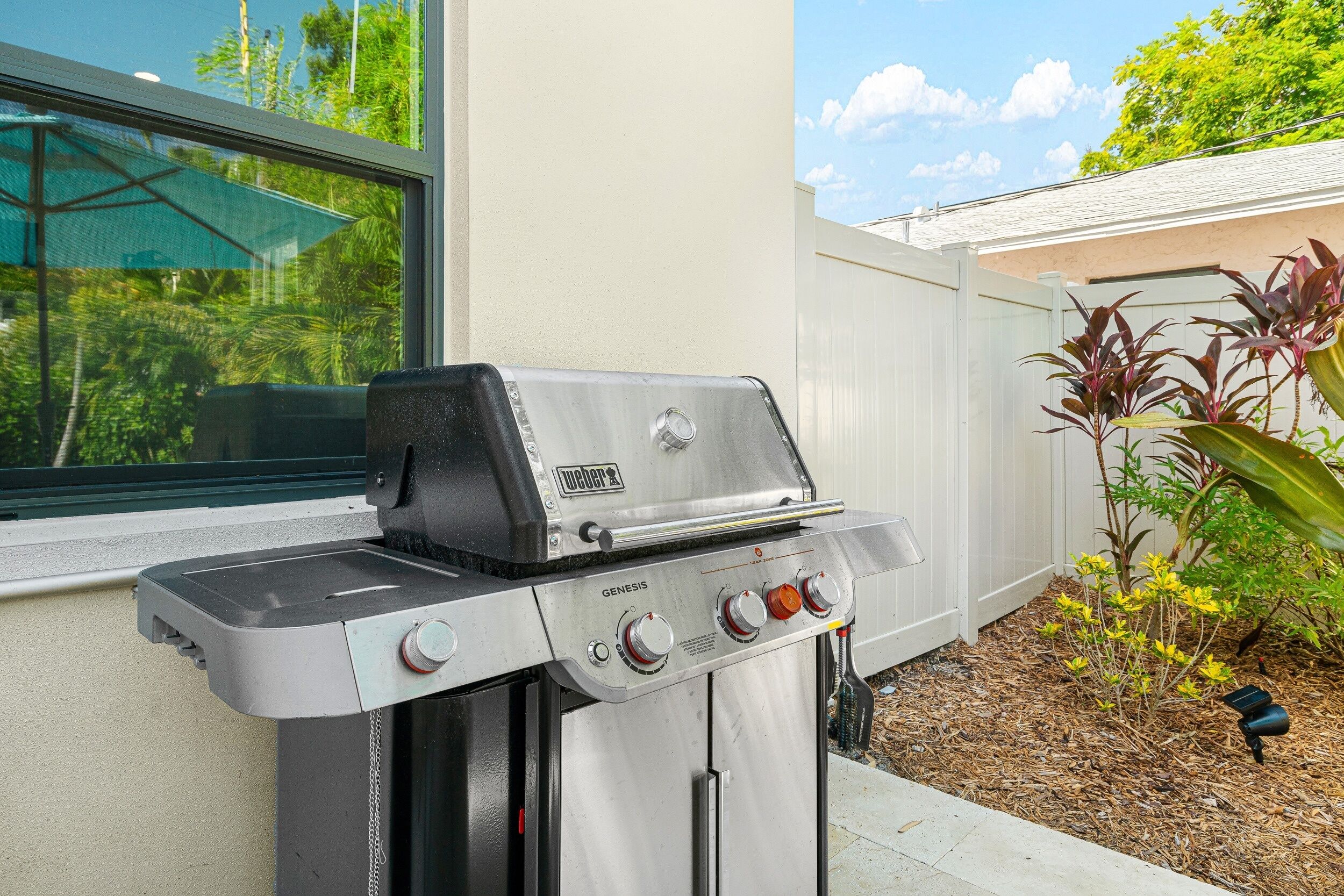 A Weber Genesis gas grill with four burners, a side shelf, and a digital display, positioned outdoors near a house with a window and a garden.