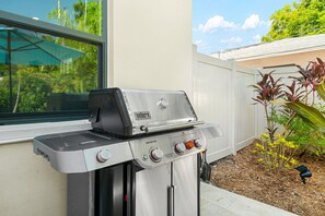 A Weber Genesis gas grill with four burners, a side shelf, and a digital display, positioned outdoors near a house with a window and a garden. - Arlington Park Oasis 4 Bedroom Home (Sarasota)