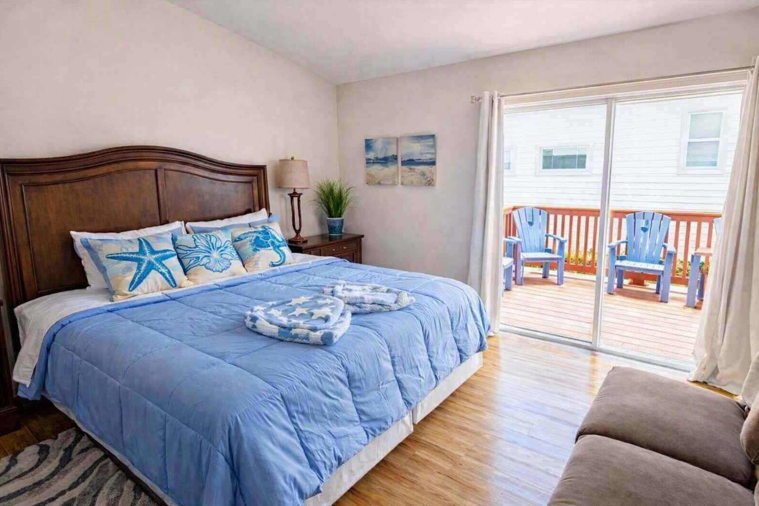 A bedroom with a large bed, a wooden headboard, a nightstand, a lamp, and a view of a deck with chairs.