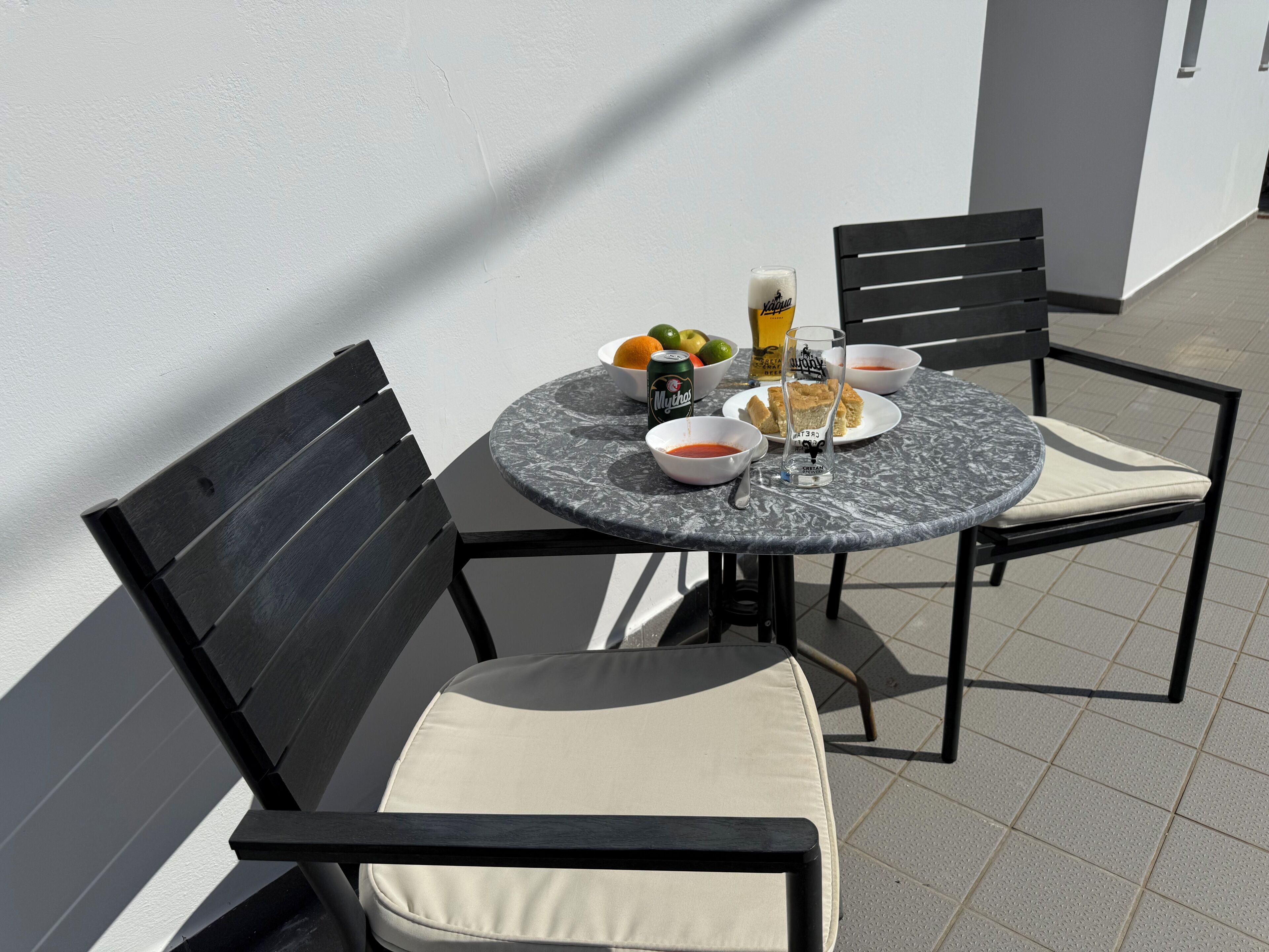 Une table ronde avec un plateau en marbre, accompagnée de deux chaises noires dotées de coussins beige. Sur la table, on trouve un verre de bière, un bol de soupe, une assiette avec une pâtisserie et une canette de bière Mythos.
